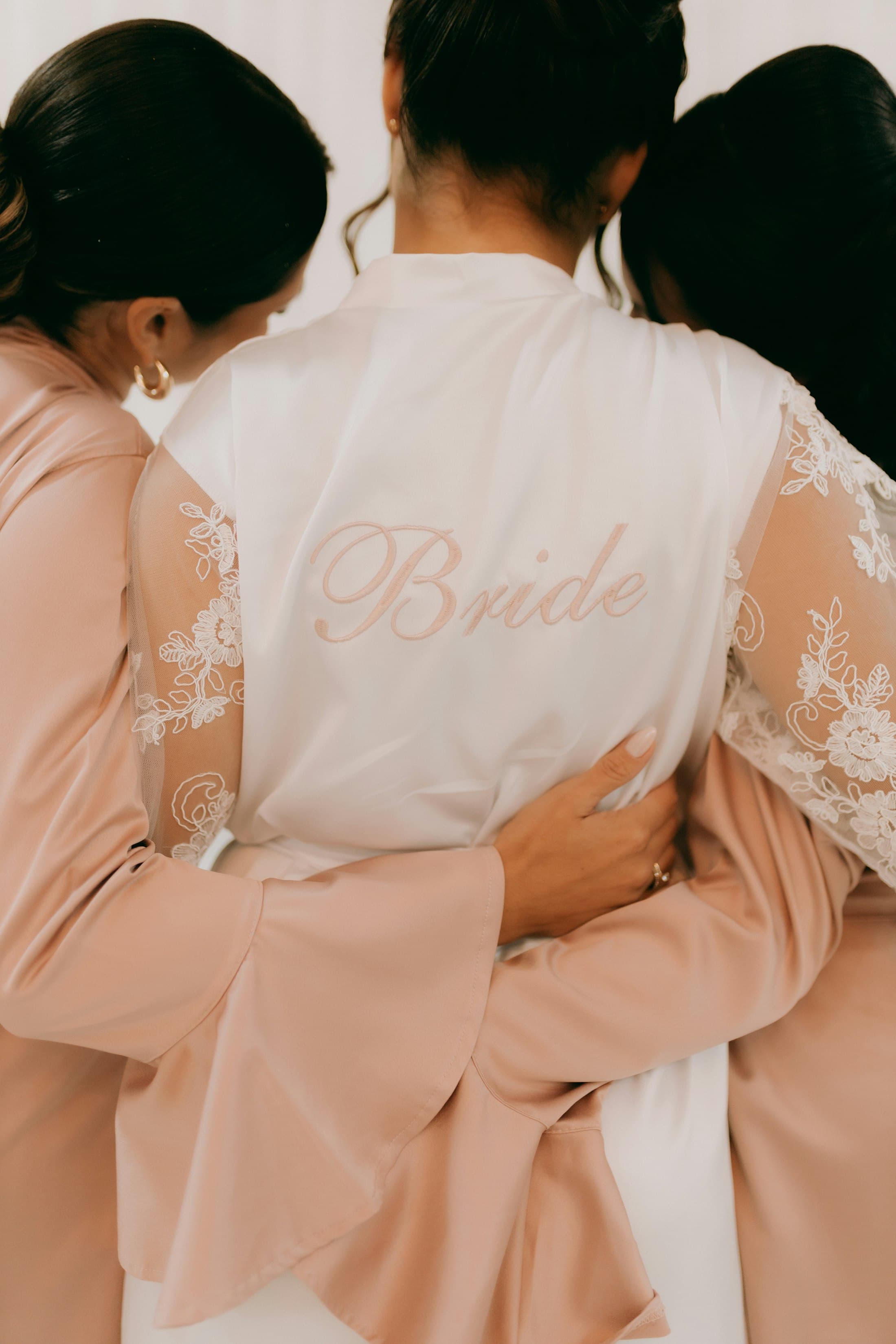 The back of a white satin robe embroidered “Bride” in blush script, two bridesmaids in blush robes holding her close.