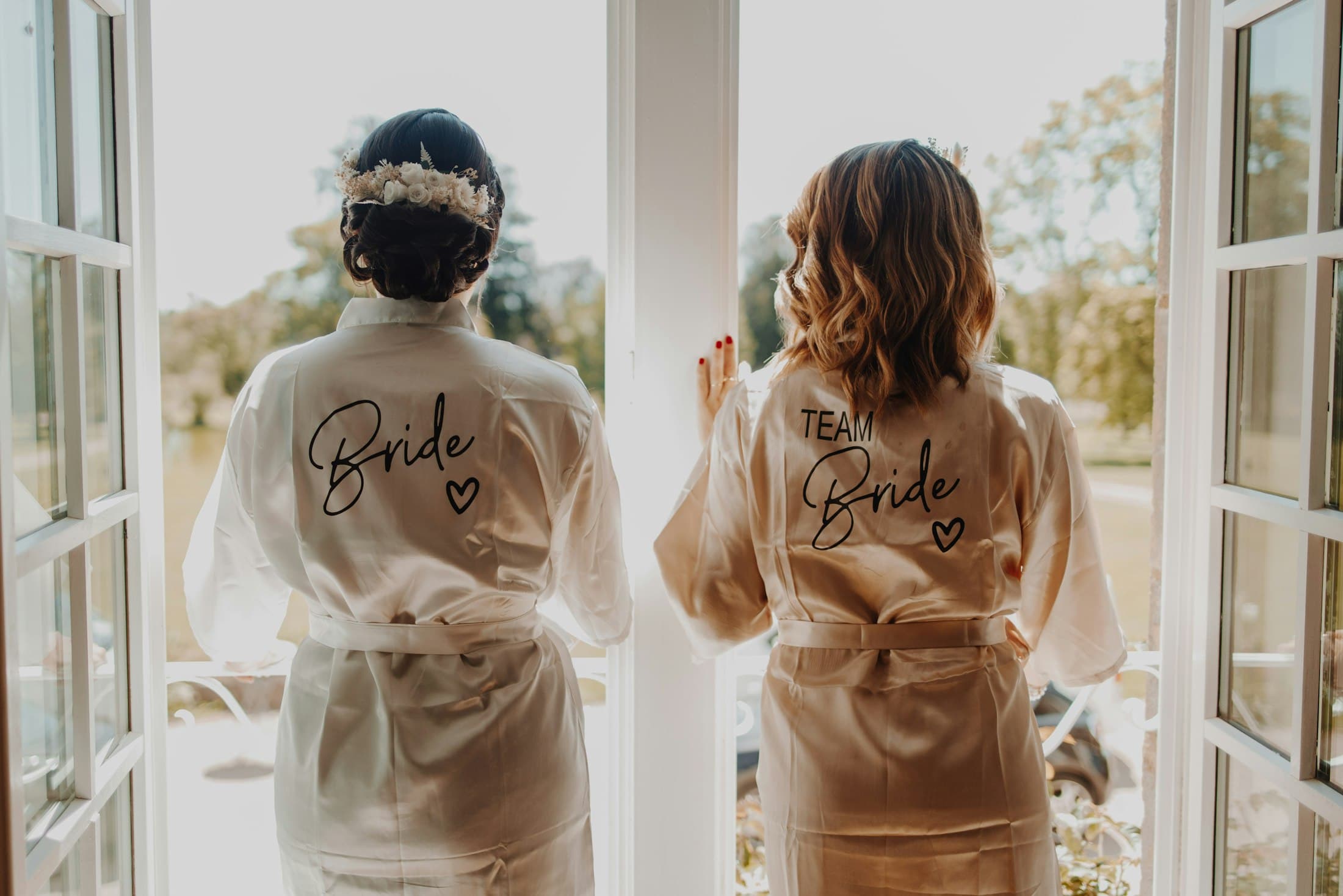 A bride and a bridesmaid at an open window in satin robes embroidered “Bride” and “Team Bride”.